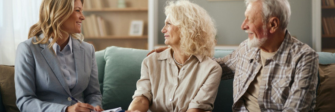 Junge Frau in Anzug sitzt mit altem Paar auf dem Sofa und schreibt auf einem Dokument