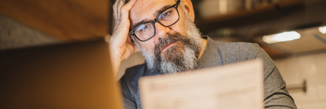 Älterer Mann sitzt am Laptop mit Dokument und stützt den Kopf in die Hand