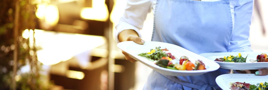 Kellnerin mit blauer Schürze trägt drei gefüllte Teller gleichzeitig.