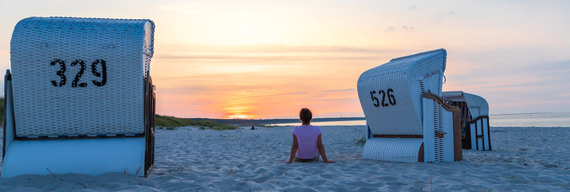 Eine Frau sitzt bei Sonnenuntergang zwischen zwei Strandkörben am Strand