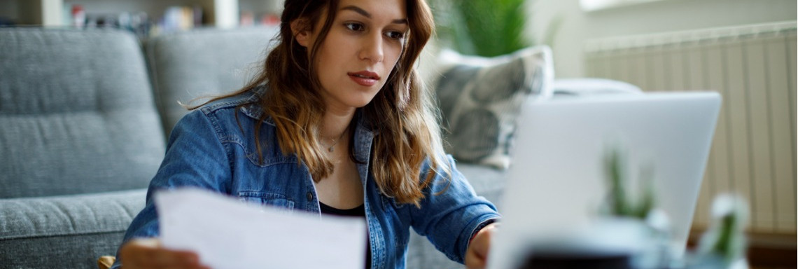 Junge Frau sitzt vor dem Sofa am Tisch und liest ein Dokument und schaut auf einen Laptop