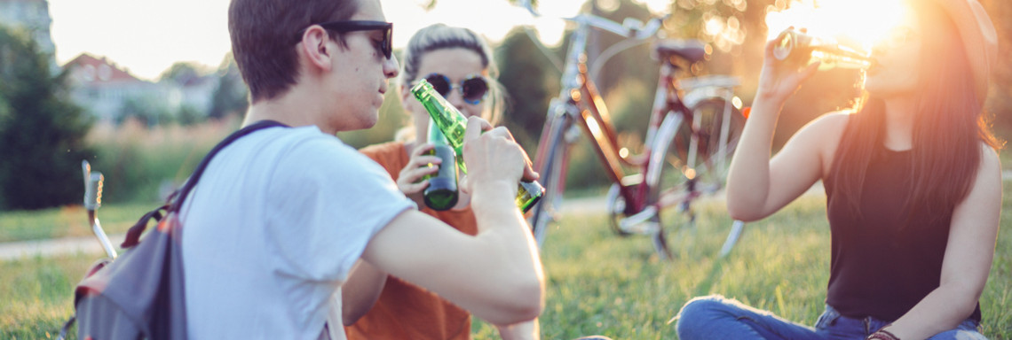 Drei junge Menschen sitzen bei Sonne mit ihren Fahrrädern auf einer Wiese und trinken Bier