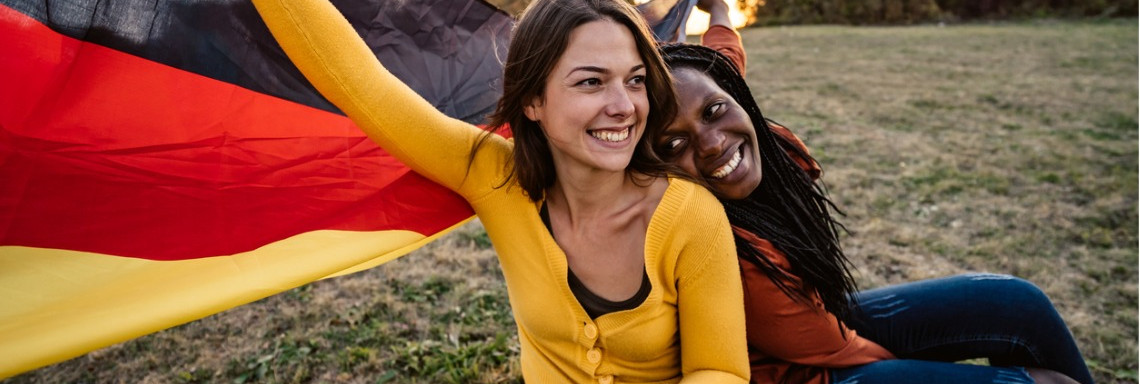 Junge weiße Frau und junge schwarze Frau sitzen mit Deutschlandflagge glücklich auf einer Wiese