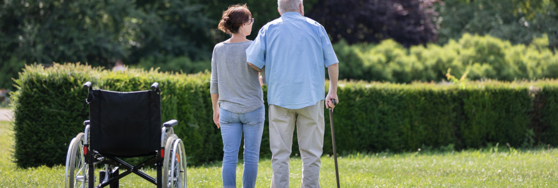 Alter Mann und Pflegerin stehen neben einem Rollstuhl in einem Garten
