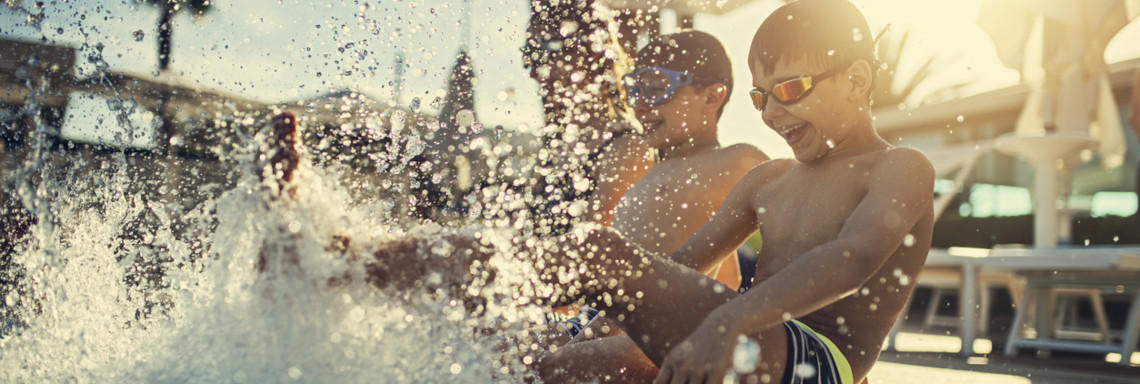 Zwei Kinder sitzen bei Sonnenschein am Beckenrand vom Pool und spritzen mit den Beinen im Wasser