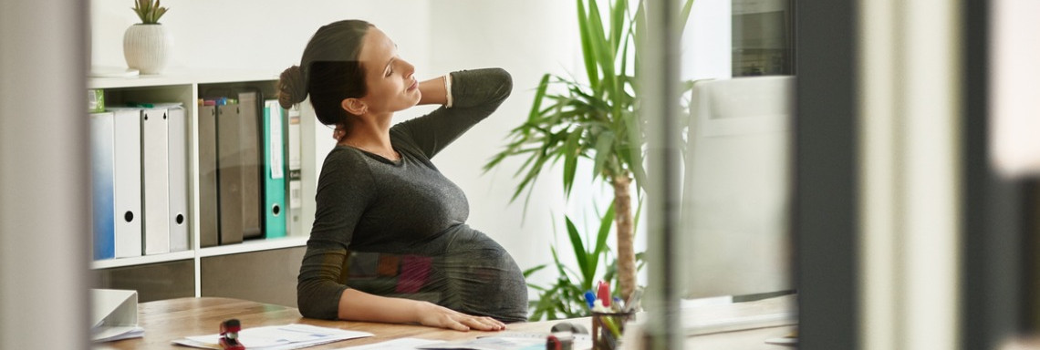 Schwangere Frau sitzt am Schreibtisch in Büro und reibt sich den Nacken