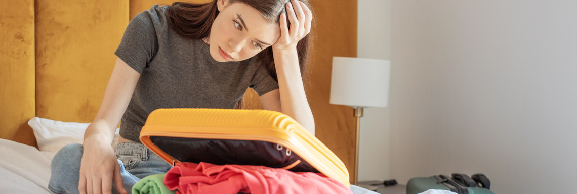 Junge Frau sitzt vor gepacktem Koffer auf Hotelbett und stützt traurig den Kopf auf die Hand