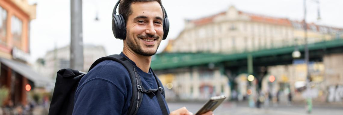 Junger Mann steht mit Kopfhörern und Handy an einer Bushaltestelle und lächelt