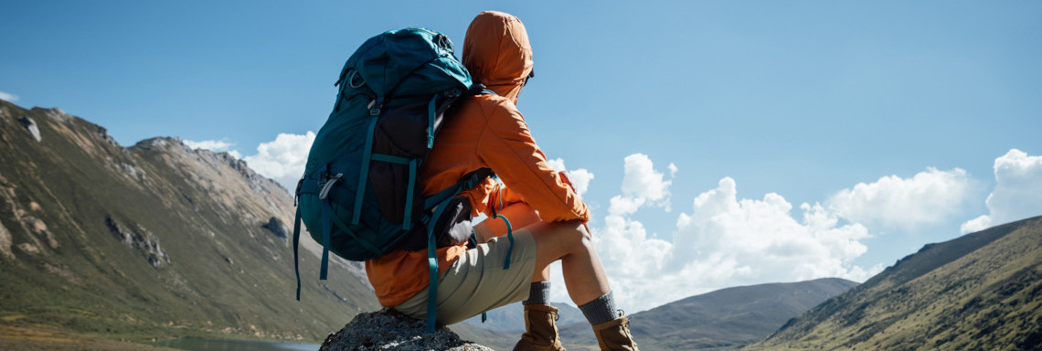 Mann in Wanderkleidung und mit Rucksack sitzt auf Stein in Gebirge und genießt Aussicht über Berge und Wasser
