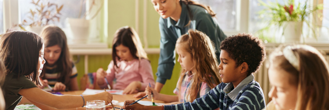 Eine Gruppe Grundschüler sitzt an einem Tisch und malt mit Wasserfarbe und die Lehrerin schaut zu