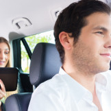 privater Taxifahrer fährt junge Frau, die mit Coffee to Go und Tablet auf dem Rücksitz sitzt