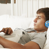 Junge mit Kopfhörern liegt im Bett, mit dem Laptop auf seinem Bauch