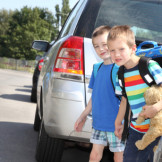 Einschulung: Schulkinder mit Ranzen und Teddbär im Arm zwischen parkenden Autos am Straßenrand
