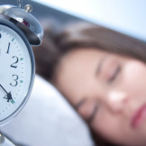 Unpünktlichkeit: Wecker steht auf 7.30 Uhr, im Hintergrund schläft eine Frau im Bett