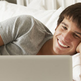 Illegale Downloads: Junger Mann liegt seitlich auf dem Bett und schaut auf seinem Laptop einen Film