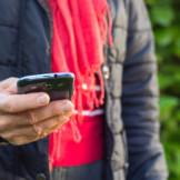 Datenautomatik beim Handytarif: Das ist zu beachten. Eine Person mit rotem Schal und dunkler Daunenjacke hält ein Smartphone in der Hand.