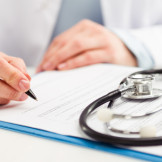 Behandlungsfehler: Besteht Anspruch auf Schmerzensgeld? Ein Arzt schreibt auf einem Klemmbrett auf dem ein Stethoskop liegt.