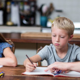 Hausaufgaben: Wie sind sie rechtlich geregelt? Ein Mann sitzt mit einem Jungen und einem Mädchen an einem Tisch und hilft bei den Hausaufgaben.