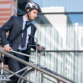 Dienstfahrrad statt Dienstwagen: Ihre Rechte als Arbeitnehmer. Ein Mann im Anzug und Fahrradhelm trägt ein Herrenrad eine kleine Treppe herunter.