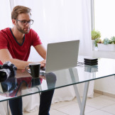 Domain anmelden: Namensschutz und Namensrecht beachten. Ein Mann mit Brille sitzt an einem gläsernen Schreibtisch an einem Laptop.