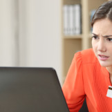 Gehalt: Verzugszinsen und Verzugspauschale fordern. Ein junge Frau in orangener Bluse sitzt mit zusammen gekniffenen Augen vor einem Laptop und hält eine Chipkarte in der Hand.