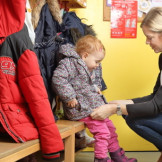 Kündigung des Kindergartenplatzes: Rechte der Eltern. Ein Frau hockt vor einem Kleinkind, das auf einer Bank sitzt, und schließt seine Jacke.