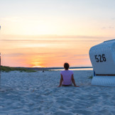 Frau sitzt im Sonnenuntergang zwischen zwei Strandkörben 