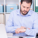 Arbeitszeitbetrug: Wann Abmahnung oder Kündigung drohen. Ein Mann in hellblauem Hemd sitzt an einem Schriebtisch im Büro und guckt auf seine Armbanduhr.
