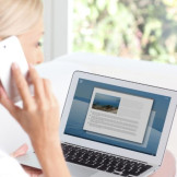 Computerkauf: Vorinstallierte Software ist zulässig. Eine blonde Frau sitzt vor einem Laptop und telefoniert mit einem Smartphone.