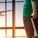 Flug übertragen: Kunde trägt Umbuchungs-Kosten. Eine Frau hält einen Rollkoffer in der Hand und schaut aus dem Fenster, in dem ein startendes Flugzeug zusehen ist.