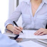 Zweitwohnsitz anmelden: Darauf müssen Sie achten. Eine Frau mit gestreifter Bluse sitzt an einem Bürotisch und zeichnet auf einem Dokument.