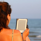 Buchpreisbindung bei eBooks: Bedeutung für Verbraucher. Eine Frau sitzt am Meer und hält einen eBook-Reader in der Hand.