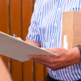 Rückgaberecht: Online-Käufer dürfen nicht unbegrenzt testen. Ein Mann in weiß-blau gestreiftem Hemd hat ein kleines Paket unterm Arm und notiert etwas auf einem Klemmbrett.