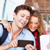 Fernreisen: Tipps zu Reisepass, Visum und Co. Ein junger Mann und eine junge Frau gucken in einem Flughafen-Terminal gemeinsam auf ein Smartphone.