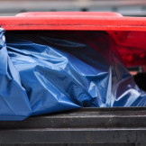 Müllentsorgung: Private Abfallpressen sind verboten. Ein graue Mülltonne mit rotem Deckel aus der ein blauer Sack ragt.