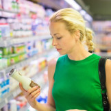 Zucker, Fett und Co.: Nährwertkennzeichnung ist Pflicht. Eine blonde Frau in grünem Kleid steht an der Kühlwarenabteilung in einem Supermarkt und hält eine kleine Milchflasche in den Händen.