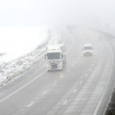 Eisplatten auf dem Lkw: Was gilt bei einem Unfall? Eine vernebelte dreispurige Autobahn, neben der Schnee liegt. Ein LKW und ein Transporter fahren auf der linken Seite.