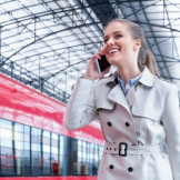 Fahrtkosten zum Bewerbungsgespräch: Wer übernimmt sie? Eine Frau im Trenchcoat telefoniert mit einem Smartphone, während sie durch ein Bahnhofsgebäude geht.
