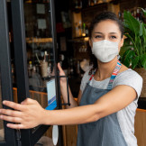 Junge Frau mit Mund-Nasen-Schutz öffnet die Tür eines Cafés