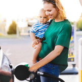 Junge Frau mit Baby auf dem Arm tankt glücklich ihr Auto