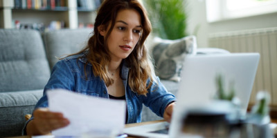 Junge Frau sitzt vor dem Sofa am Tisch und liest ein Dokument und schaut auf einen Laptop