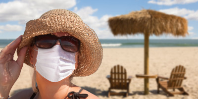Frau mit Sonnenbrille und medizinischer Maske sitzt am Strand Frau mit Sonnenbrille und medizinischer Maske sitzt am Strand