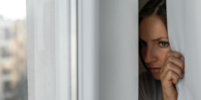 Frau schaut verängstigt hinter Gardine durchs Fenster