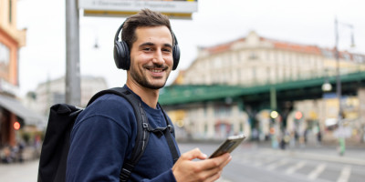 Junger Mann steht mit Kopfhörern und Handy an einer Bushaltestelle und lächelt