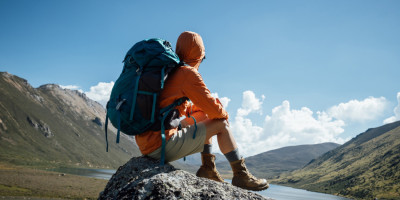 Mann in Wanderkleidung und mit Rucksack sitzt auf Stein in Gebirge und genießt Aussicht über Berge und Wasser