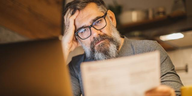 Älterer Mann sitzt am Laptop mit Dokument und stützt den Kopf in die Hand