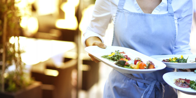 Kellnerin mit blauer Schürze trägt drei gefüllte Teller gleichzeitig.