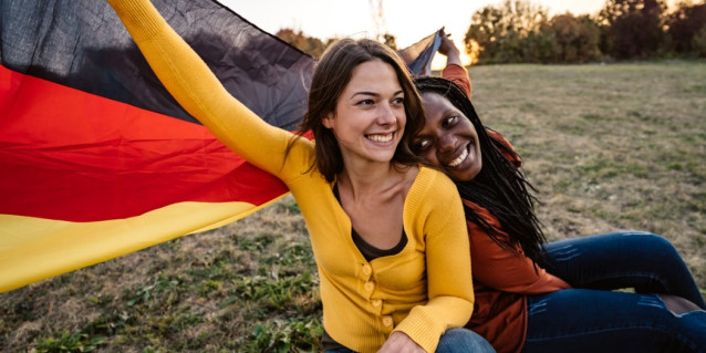 Junge weiße Frau und junge schwarze Frau sitzen mit Deutschlandflagge glücklich auf einer Wiese