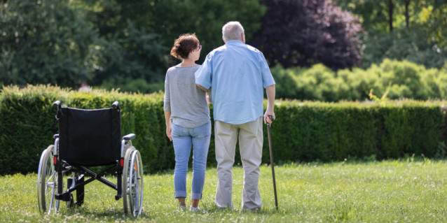 Alter Mann und Pflegerin stehen neben einem Rollstuhl in einem Garten