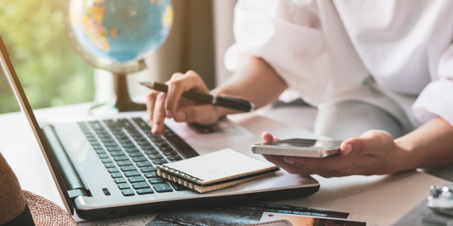 Person macht Urlaubsplanung mit Laptop, Smartphone und Notizblock